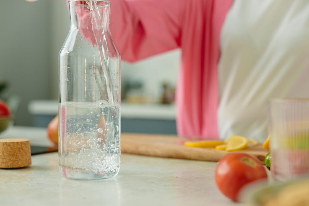 A fresh glass pitcher being filled with water, surrounded by lemon slices and tomatoes on a kitchen counter.