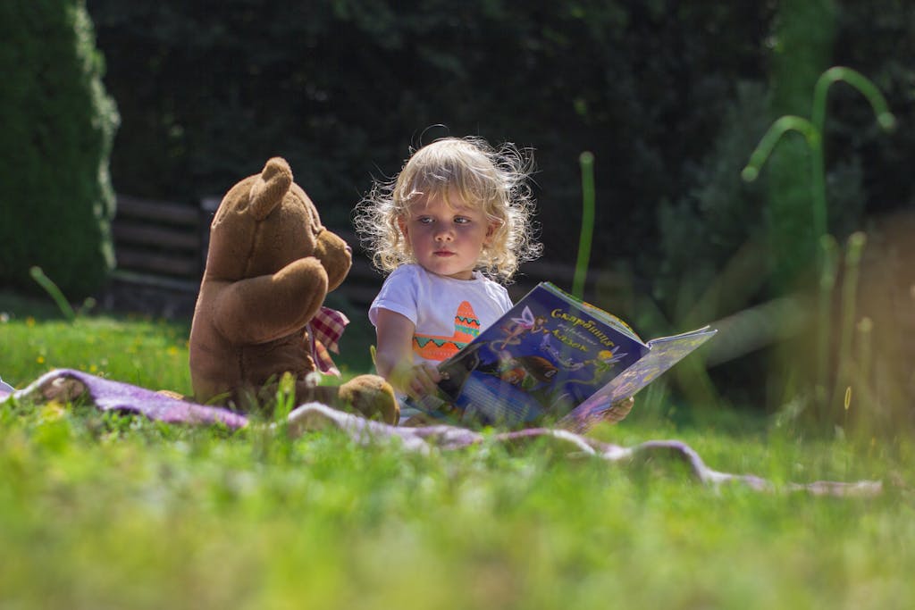 Young child reading a book outdoors with a teddy bear, enjoying a sunny day on the grass.
